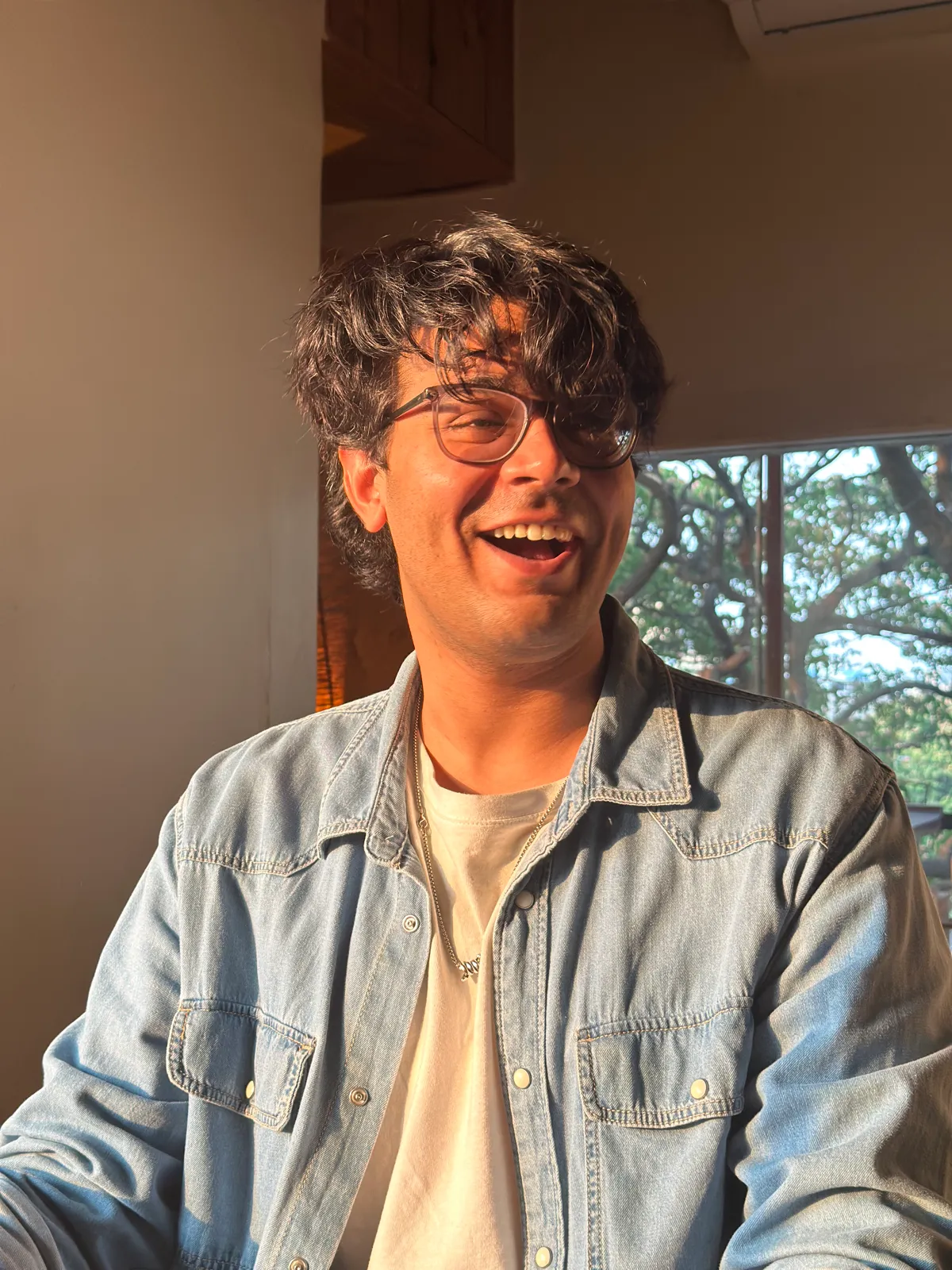 Hishaam laughing, in glasses and a denim shirt, in warm afternoon light